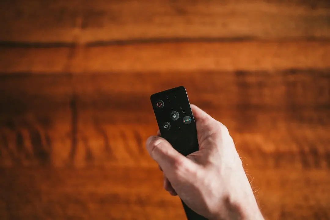 Hand pointing a small black TV remote control against a warm wooden background