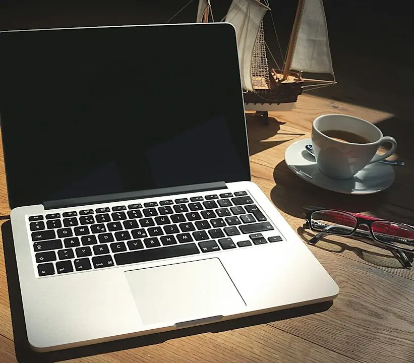 Open laptop on a wooden desk next to a cup of coffee, reading glasses and a model sailing ship
